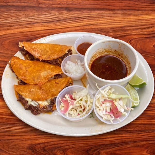 Birria tacos with consommé, chopped onions, coleslaw, and lime wedges on a white plate.