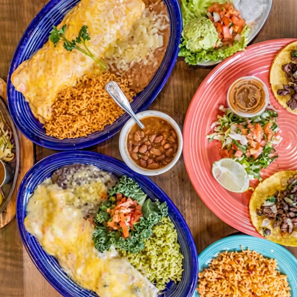 Colorful spread of Mexican dishes: cheesy burrito, steak fajitas, street tacos, rice, beans, and guacamole.