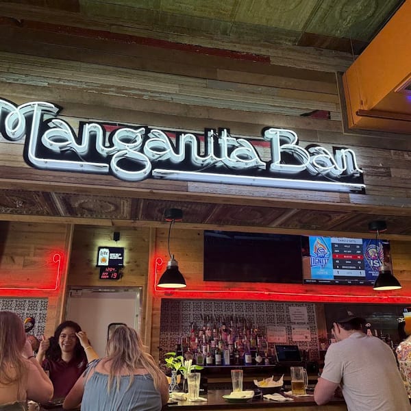 A neon "Margarita Bar" sign illuminates a lively bar with patrons and a bartender.